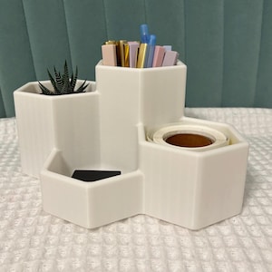 May include: White hexagonal desk organizer with four compartments. The top compartment holds pens and pencils in various colors. The middle compartment holds a small succulent plant. The bottom compartment holds a roll of tape. The organizer is made of white plastic.