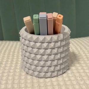 May include: A gray, textured pencil holder with a studded design, filled with pastel-colored highlighters. The highlighters are in shades of beige, green, blue, pink, and orange. The holder sits on a white, textured surface.