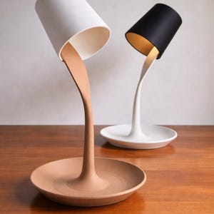 以下が含まれることがあります： ユニークなデザインのモダンなテーブルランプ2つ。1つのランプは白いシェードとタン色のベース、もう1つは黒いシェードと白いベースです。ランプは液体を注いでいるように見え、芸術的な効果を生み出しています。