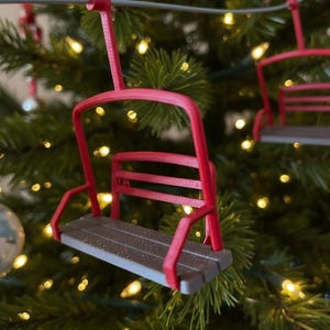 May include: A red ski lift Christmas tree decoration with a brown seat and a red frame. The ornament is suspended on a Christmas tree with fairy lights. The ski lift has a detailed design.
