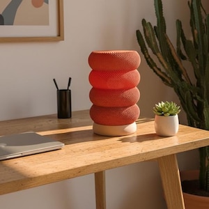 May include: A modern orange cylindrical lamp with a textured surface, composed of stacked rings, sits on a wooden desk. A small succulent in a white pot and a black pen holder are also on the desk. A laptop is partially visible.