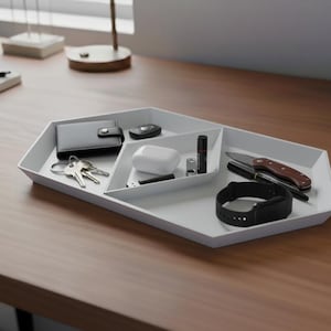 Può includere: Un vassoio organizzatore da scrivania bianco e geometrico con più scomparti. Il vassoio contiene un portafoglio nero, chiavi, un telecomando per auto, auricolari wireless, una piccola torcia, una penna, un coltello tascabile e uno smartwatch nero.