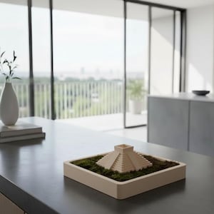 Puede incluir: Una escultura de pirámide en miniatura beige se encuentra en una jardinera rectangular llena de musgo verde. La jardinera está sobre una encimera oscura. Un jarrón blanco con vegetación y una pila de libros también están en la encimera.