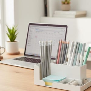May include: A white desk organizer filled with pencils, pens, and sticky notes sits on a light wood desk. A laptop, coffee mug, glasses, and a small potted plant are also on the desk, creating a workspace.