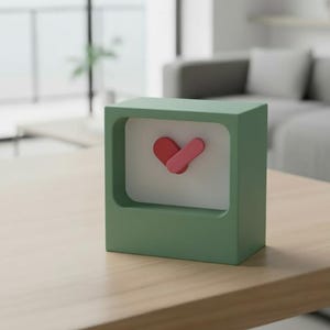 Puede incluir: Un reloj verde en forma de cubo con una esfera blanca y un diseño en forma de corazón rojo y rosa. El reloj está sobre una mesa de madera. El fondo incluye un sofá y plantas.