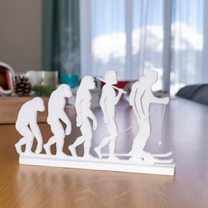 May include: A white decorative sculpture depicting the evolution of man, culminating in a skier. The sculpture is placed on a wooden table. Ski goggles and other decorative items are in the background.