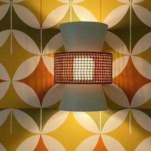 May include: A retro wall light with a light blue top and bottom, and a brown, grid-patterned centre. It's mounted on wallpaper with a repeating geometric pattern in yellow, orange, green, and white. The light is on.