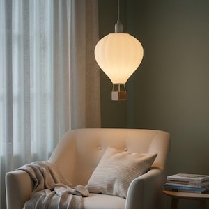 May include: A white hot air balloon-shaped pendant light hangs above a cream armchair. The light has a wooden basket and is suspended from the ceiling. A throw and pillow rest on the chair. The room has a window with white curtains.
