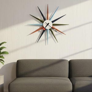 May include: A starburst-style wall clock with colorful, pointed arms in shades of orange, blue, green, and black. The clock face is white with black hands. A potted plant sits on a small, round, gray table next to a gray sofa.