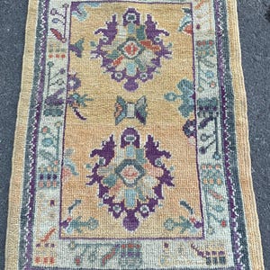 May include: A vintage Turkish rug with a yellow background and a purple, green, and blue floral pattern. The rug has a fringe border.