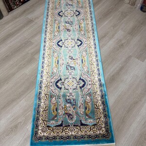 May include: A rectangular, turquoise runner rug with an intricate paisley and floral pattern in cream, navy, and yellow. The rug has a turquoise border and cream fringe on both ends. The rug is placed on a light wood floor.