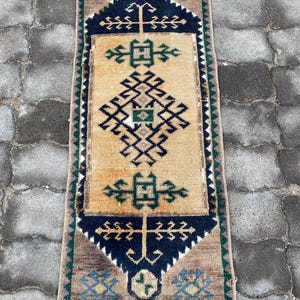 Puede incluir: Una alfombra rectangular de estilo vintage con un campo central beige y intrincados patrones geométricos en azul marino, verde y marrón. La alfombra presenta un borde con motivos florales repetidos y un borde con flecos. Mide aproximadamente 60 cm de ancho y 122 cm de largo.