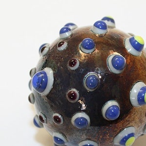 May include: A handcrafted glass orb with a textured, brown surface. The orb is adorned with numerous blue, white, and yellow eye-like protrusions. The text "Brad Pearson" and "1 1/2" are visible.