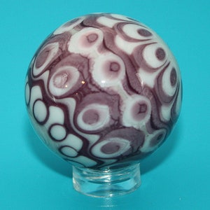 May include: A decorative glass sphere with a swirling pattern of white, lavender, and dark purple. The sphere is resting on a clear, round base, and the background is a solid teal color.