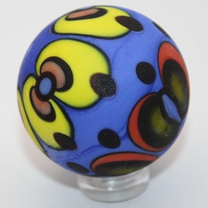 May include: A colorful glass marble with a blue base, adorned with yellow, red, and black abstract designs. The marble is round and sits on a clear, rounded stand. The design is reminiscent of a flower.