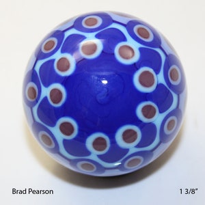 May include: A blue glass marble with a white and purple swirl pattern. The marble is 1 3/8 inches in diameter.