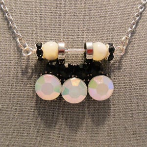 May include: A silver necklace featuring a pendant with a barbell design. The pendant has cream-colored beads, black beads, and three iridescent, round, clear gemstones. The necklace is on a gray background.