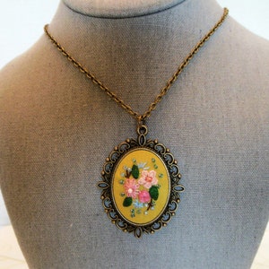 Może przedstawiać: Złoty łańcuszek z ż&oacute;łtym owalnym wisiorkiem, na kt&oacute;rym wyszyto r&oacute;żowe i białe kwiaty z zielonymi liśćmi. Wisiorek jest ozdobiony dekoracyjną złotą metalową ramką.