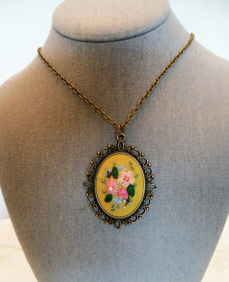 Może przedstawiać: Złoty łańcuszek z ż&oacute;łtym owalnym wisiorkiem, na kt&oacute;rym wyszyto r&oacute;żowe i białe kwiaty z zielonymi liśćmi. Wisiorek jest ozdobiony dekoracyjną złotą metalową ramką.