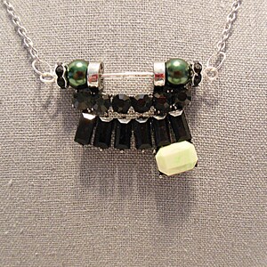 May include: A silver-toned necklace featuring a pendant with black and green accents. The pendant has a central bar with green beads on each end, black rectangular and round stones, and a light green rectangular stone.