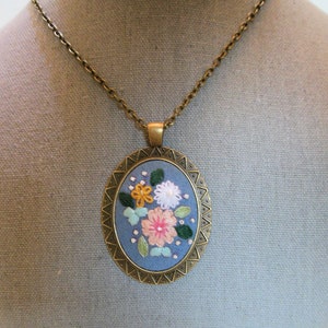May include: A blue and gold oval pendant necklace with a floral embroidery design. The flowers are pink, white, and yellow.