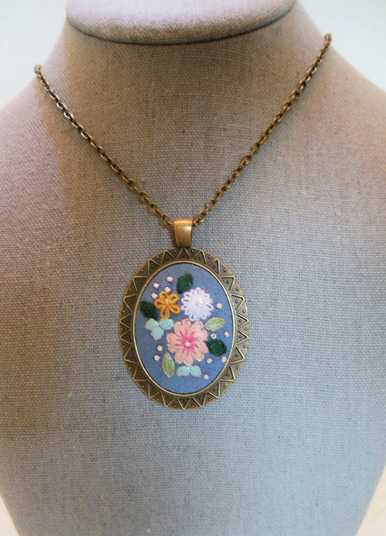 May include: A blue and gold oval pendant necklace with a floral embroidery design. The flowers are pink, white, and yellow.