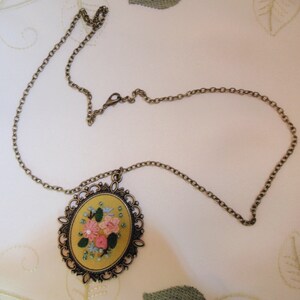 Może przedstawiać: Złoty łańcuszek z owalnym wisiorkiem z haftem kwiatowym. Haft wykonany jest nicią w kolorze r&oacute;żowym, zielonym i niebieskim na ż&oacute;łtym tle z tkaniny.