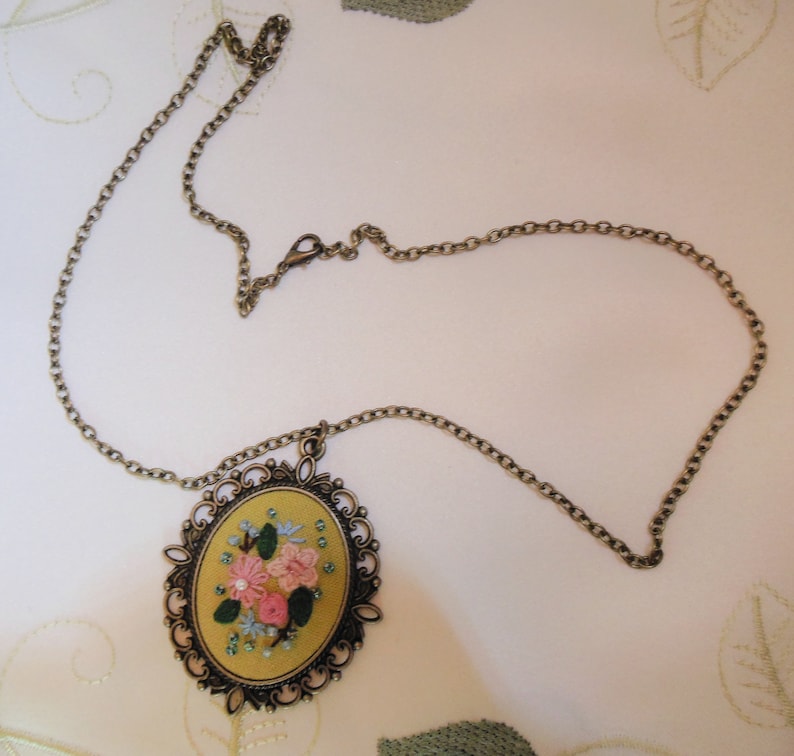 Może przedstawiać: Złoty łańcuszek z owalnym wisiorkiem z haftem kwiatowym. Haft wykonany jest nicią w kolorze r&oacute;żowym, zielonym i niebieskim na ż&oacute;łtym tle z tkaniny.