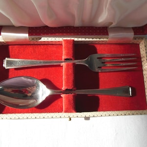 May include: A silver baby spoon and fork set in a red velvet lined box. The set is in a white box with a black and white pattern.
