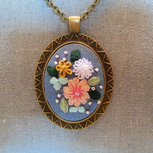 May include: A bronze pendant necklace with a blue fabric circle featuring embroidered flowers in pink, yellow, and white.