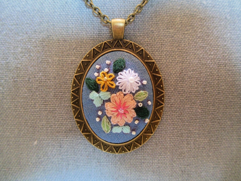 May include: A bronze pendant necklace with a blue fabric circle featuring embroidered flowers in pink, yellow, and white.