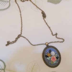 May include: A delicate gold chain necklace with a round pendant featuring a blue fabric background with embroidered flowers in shades of pink, yellow, orange, and white.