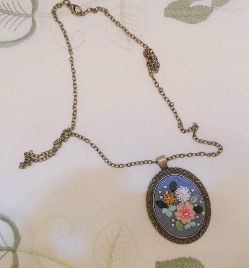 May include: A delicate gold chain necklace with a round pendant featuring a blue fabric background with embroidered flowers in shades of pink, yellow, orange, and white.