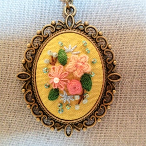 Może przedstawiać: Ż&oacute;łty haft kwiatowy w brązowej owalnej ramie. Wz&oacute;r przedstawia r&oacute;żowe i niebieskie kwiaty z zielonymi liśćmi i jasnoniebieskimi koralikami.