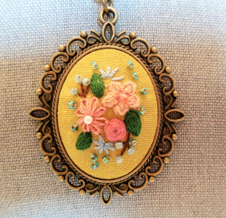 Może przedstawiać: Ż&oacute;łty haft kwiatowy w brązowej owalnej ramie. Wz&oacute;r przedstawia r&oacute;żowe i niebieskie kwiaty z zielonymi liśćmi i jasnoniebieskimi koralikami.