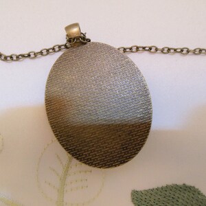 May include: A bronze oval pendant with a textured surface hangs from a bronze chain. The pendant is about 2 inches in diameter.