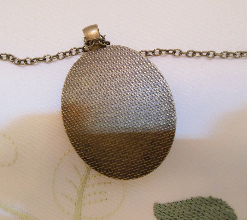 May include: A bronze oval pendant with a textured surface hangs from a bronze chain. The pendant is about 2 inches in diameter.