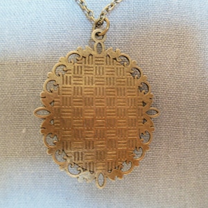 Może przedstawiać: Złoty naszyjnik z wisiorem w kształcie owalu, ozdobiony dekoracyjnym wzorem filigranowym. Wisiorek ma fakturowaną powierzchnię.