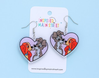 May include: Heart-shaped dangle earrings featuring a cartoon image of two dogs kissing. The earrings have a lavender background and are attached to a white card with the text "Inspired by Main Street". The earrings are on a light blue background.