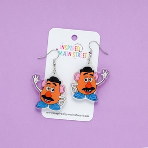 May include: A pair of novelty earrings featuring a cartoon character with an orange face, black mustache, and blue shoes. The earrings are on a white card with the text "INSPIRED BY MAIN STREET" and a website address.
