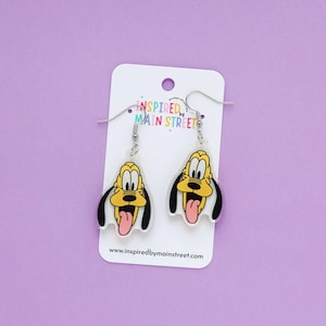 May include: A pair of dangle earrings featuring a cartoon dog face with yellow, black, and pink accents. The earrings are attached to a white card with the text "INSPIRED BY MAIN STREET" and the website address www.inspiredbymainstreet.com.