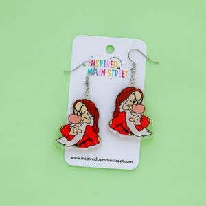 May include: A pair of dangle earrings featuring a cartoon character in a red coat and hat, with a white beard and a grumpy expression. The earrings are on a white card with the text "Inspired by Main Street".