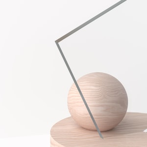 May include: A light-colored wooden sphere rests on a matching circular platform. A geometric, gray-lined shape intersects the sphere and platform, creating a minimalist, abstract composition against a white background.