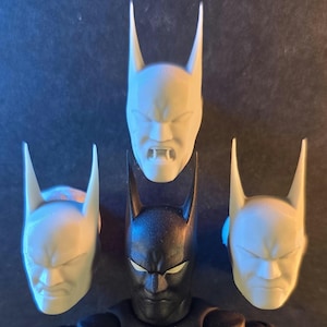 May include: A black and red Batman action figure with four interchangeable white Batman head sculpts. The figure has a stern expression and is posed against a dark background. The head sculpts have different expressions.