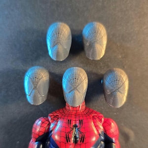 May include: A Spider-Man action figure with interchangeable heads. The figure has a red and black suit with a spider emblem. Five gray Spider-Man mask heads are displayed, each with a web pattern.