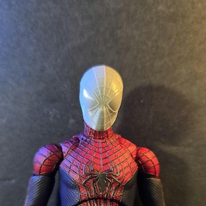 May include: A detailed action figure of Spider-Man, featuring a red and black suit with a spider emblem on the chest. The figure's mask is half white and half gray, with web detailing. The figure is posed against a dark background.