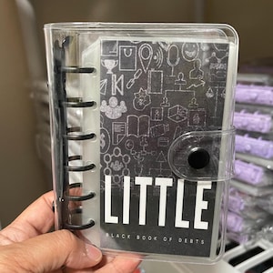 May include: A clear plastic binder with a black cover that says "LITTLE BLACK BOOK OF DEBTS". The cover has a black and white pattern of various icons and symbols.