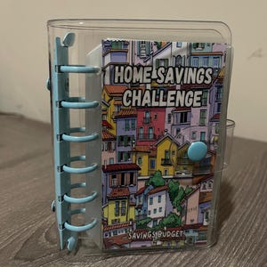 Pode incluir: Uma pasta de plástico transparente com lombada azul clara e fecho de botão. A pasta contém um insert colorido com o texto "HOME SAVINGS CHALLENGE" e um desenho de edifícios. A parte inferior do insert diz "SAVINGS BUDGET".