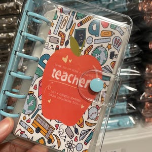 May include: A clear plastic binder with a red apple design and the text "Thank you for being a wonderful teacher. Just a friendly reminder during challenging days!" The apple has a blue button on it.