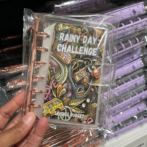 May include: A clear plastic binder with a colorful doodle design. The cover features the text "Rainy Day Challenge" and "Savings Budget".